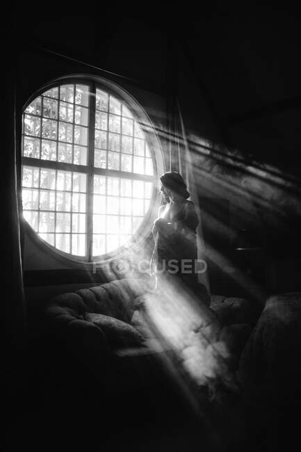Bianco e nero donna irriconoscibile in abito in piedi sul divano contro finestra di forma rotonda in casa nella giornata di sole — Foto stock
