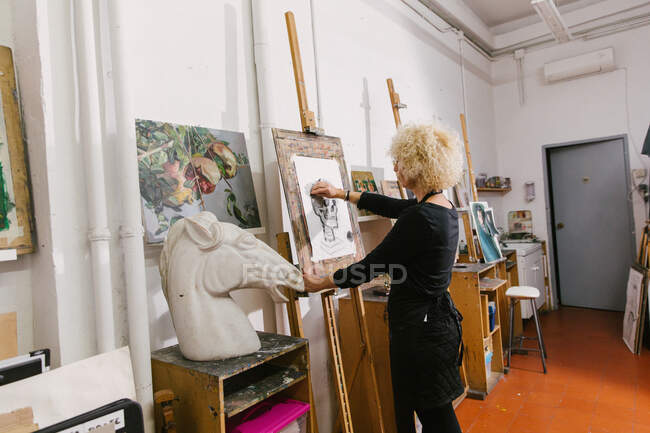Vista lateral de talentosa artista femenina de pie en el caballete y dibujo sobre papel con lápiz en el estudio de arte - foto de stock