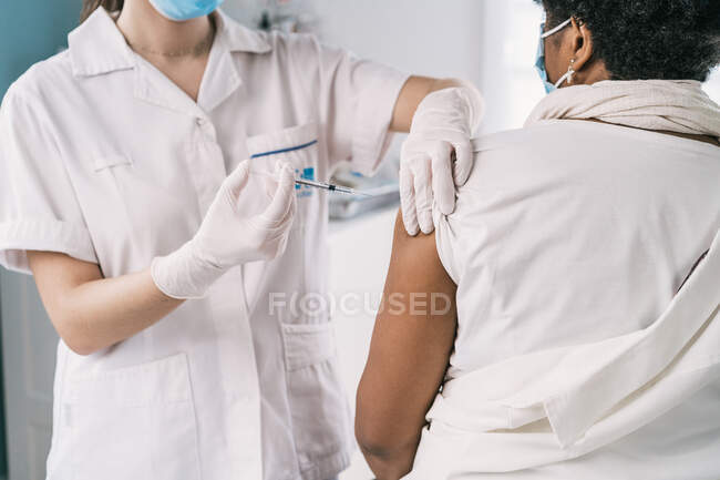 Especialista médica femenina irreconocible en uniforme protector, guantes de látex y mascarilla facial vacunando a una paciente madura afroamericana anónima en la clínica durante el brote de coronavirus - foto de stock