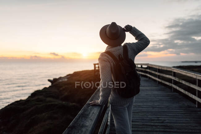 Man with backpack and hat standing on a walkway looking at the sea — Stock Photo