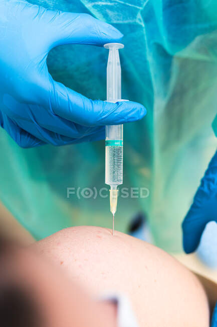 Especialista médica femenina en uniforme protector, guantes de látex y mascarilla facial vacunando a un paciente hispano en clínica durante un brote de coronavirus - foto de stock