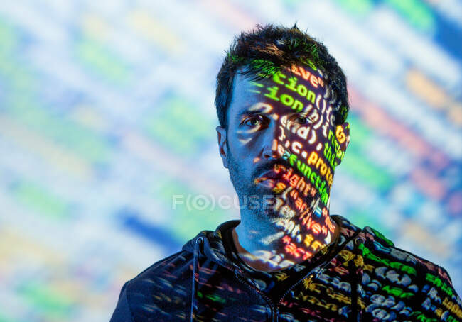 Adult bearded male in hoodie with multicolored words on half of face from projector light looking at camera — Stock Photo