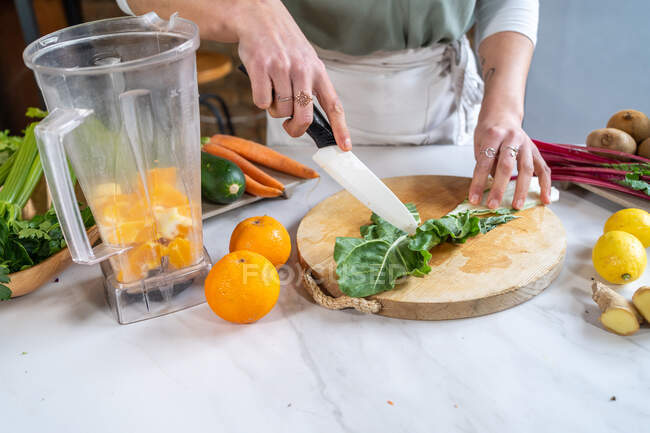 Corte feminino irreconhecível cortando folhas de acelga frescas na tábua de corte contra tigela de liquidificador com fatias de laranja na cozinha da casa — Fotografia de Stock
