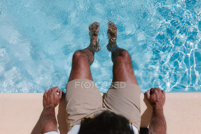 Vista superior de un freelancer masculino irreconocible sentado junto a la piscina con las piernas en el agua durante el teletrabajo en verano - foto de stock