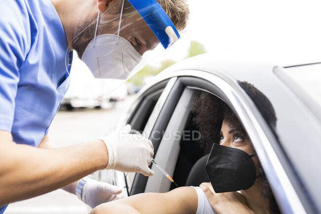 Seitenansicht eines Arztes in Schutzuniform, Latexhandschuhen und Gesichtsschutz, der eine afroamerikanische Patientin während einer Fahrt durch eine mobile Klinik während des Coronavirus-Ausbruchs im Auto impft — Stockfoto