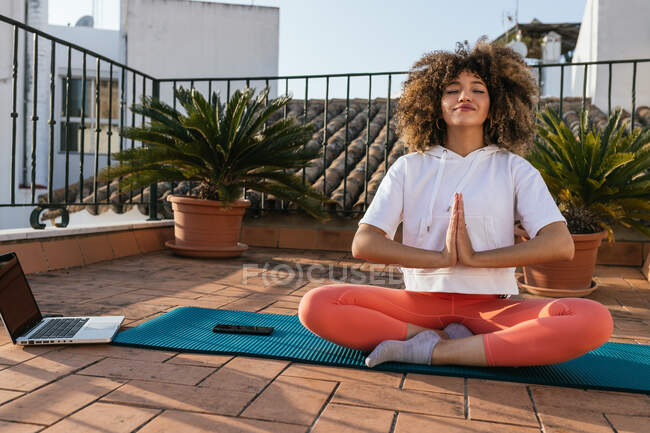 Tranquilo afro-americano feminino sentado em Padmasana com gesto Namaste e meditando com os olhos fechados enquanto pratica ioga no telhado — Fotografia de Stock