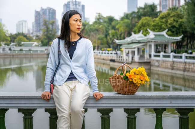 Hermosa chica asiática retrato en un parque mientras ella se sienta al lado de canasta de mimbre con flores amarillas. - foto de stock
