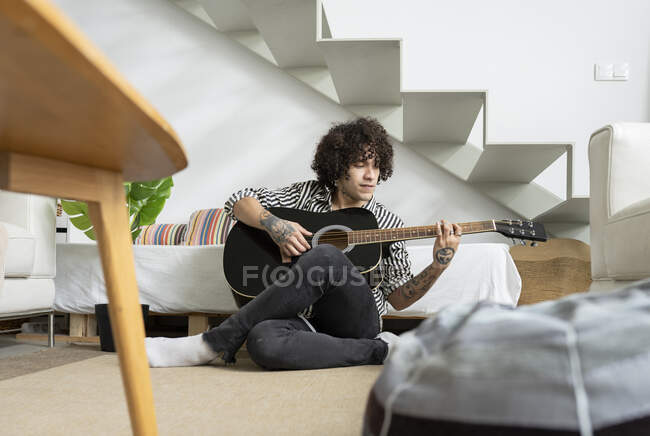 Jeune guitariste masculin tatoué assis sur le sol et jouant de la guitare à la maison — Photo de stock