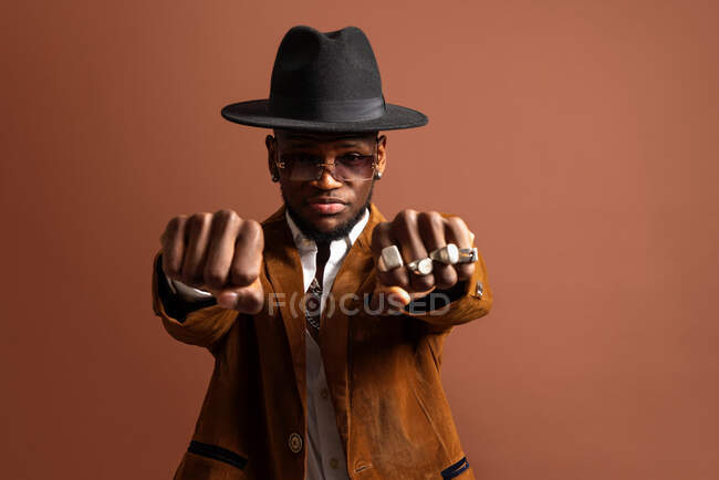 Молодий етнічний чоловік в капелюсі і стильний одяг дивиться на камеру на коричневому фоні — стокове фото