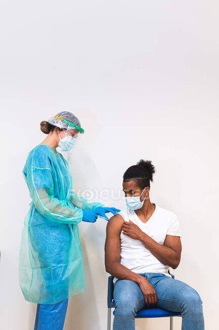 Специалистка-женщина в защитной форме, латексных перчатках и маске для лица, прививающая больного афроамериканца в клинике во время вспышки коронавируса — стоковое фото