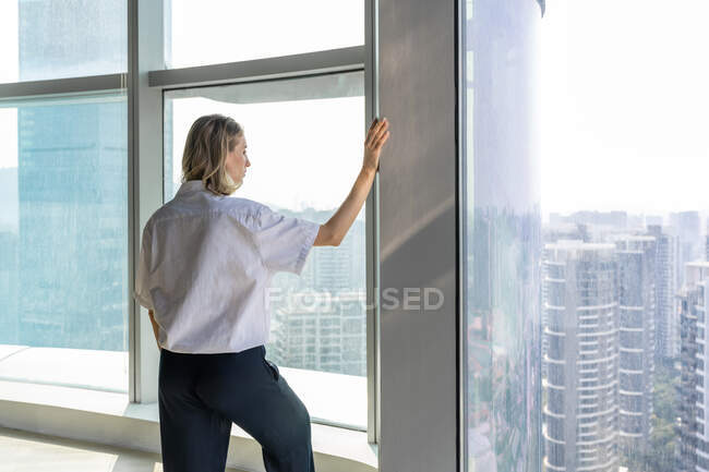 Вид ззаду на самотню дівчину в порожньому офісі, спостерігаючи за містом з великого вікна — стокове фото