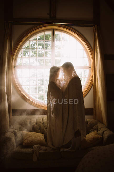 Vista lateral da namorada irreconhecível com macho amado sob tecido abraçando no sofá contra a janela em forma redonda na luz solar — Fotografia de Stock