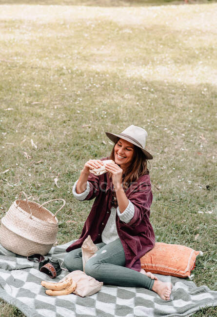 Feminino em desgaste casual e chapéu desfrutando de saboroso sanduíche enquanto sentado em tecido perto de cesta de vime e olhando para longe — Fotografia de Stock