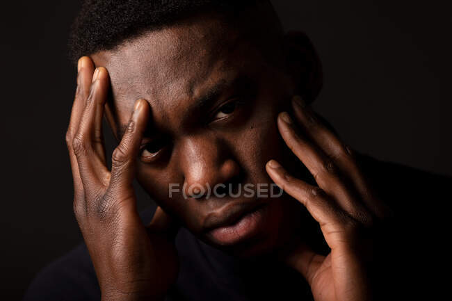 Crop unglücklichen jungen schwarzen Mann in schwarzen Pullover mit den Händen auf dem Gesicht Blick auf die Kamera auf schwarzem Hintergrund in hellen Studio — Stockfoto