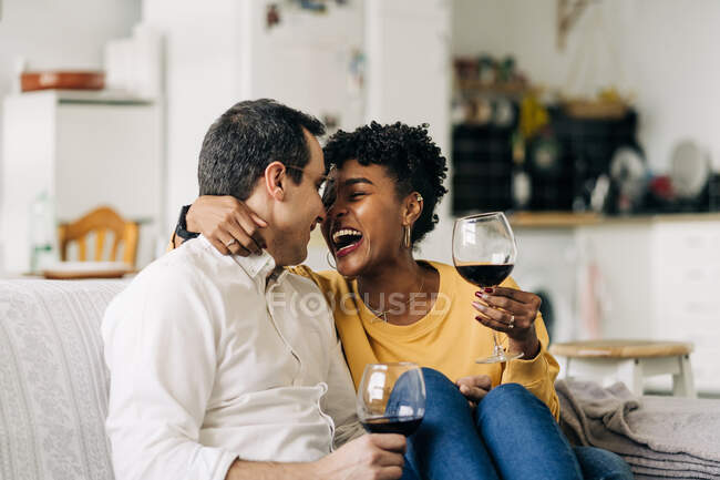 Вміст багаторасової пари охолоджує диван вдома з червоним вином в окулярах, насолоджуючись вихідними вдома і дивлячись один на одного — стокове фото