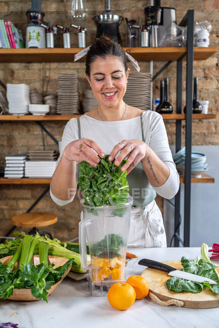 Female putting fresh chard leaves blender bowl with orange slices in house kitchen — Stock Photo