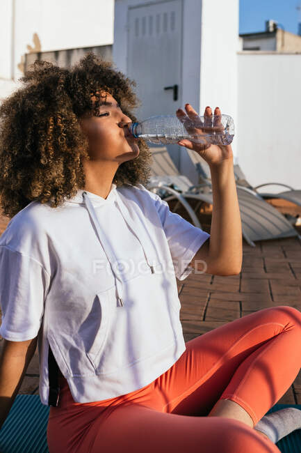 Вид сбоку чернокожей женщины в костюме, сидящей на крыше и пьющей воду после урока йоги — стоковое фото