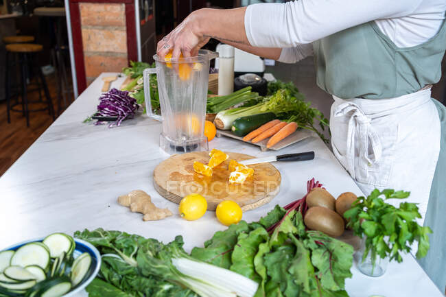 Вид сбоку на неузнаваемый урожай женщины кладет свежие оранжевые ломтики в миску блендера за столом с разнообразными овощами на кухне — стоковое фото