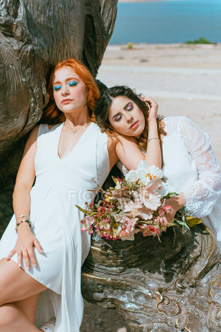 Giovane sposa in abito bianco che abbraccia femminile amato vicino mazzo di fiori sulla costa durante l'evento festivo — Foto stock
