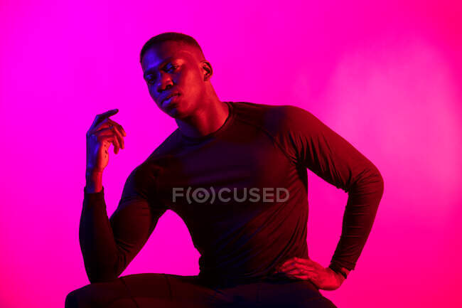 Уверенный молодой черный мужчина в темном спортивном наряде смотрит на камеру на неоновом розовом фоне в темной студии — стоковое фото