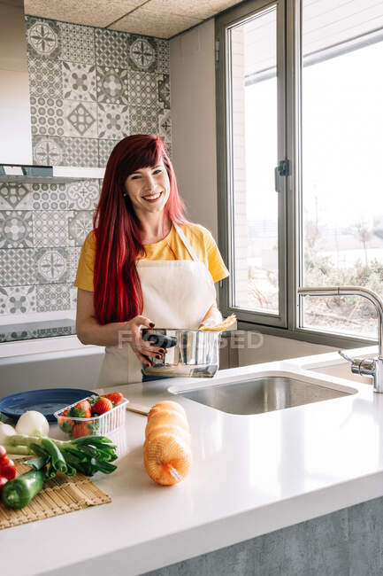 Young homosexual female pouring water from tap into saucepan with uncooked pasta against assorted vegetables in house — Stock Photo