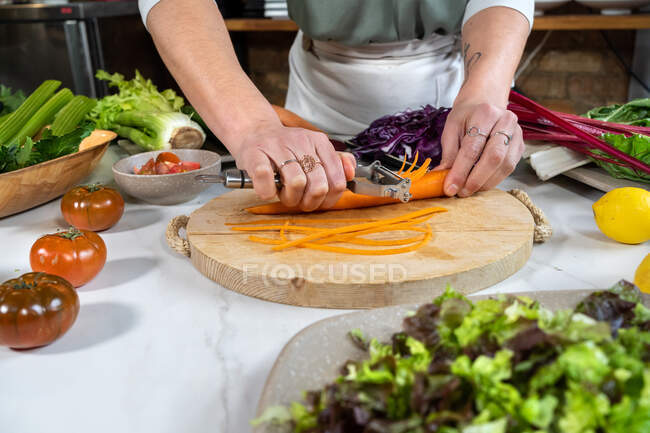Ernte bis zur Unkenntlichkeit Weibchen schneiden rohe Karotte mit Schäler bei der Zubereitung von vegetarischem Essen im Haus — Stockfoto
