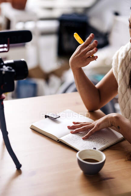 Обрізаний невпізнаваний жіночий відеоблогер з блокнотом, що сидить за столом з фотоапаратом на тринозі на кухні — стокове фото