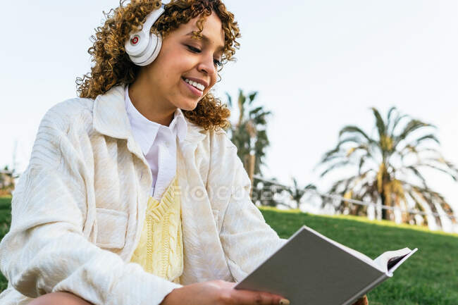Baixo ângulo de feliz afro-americano fêmea em fones de ouvido sentado no parque exótico e lendo livro interessante no verão — Fotografia de Stock