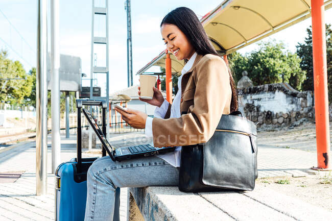 Вид сбоку счастливой азиатской женщины-фрилансера, сидящей на лавочке с ноутбуком и просматривающей смартфон в ожидании поезда на вокзале и спокойно работающей — стоковое фото