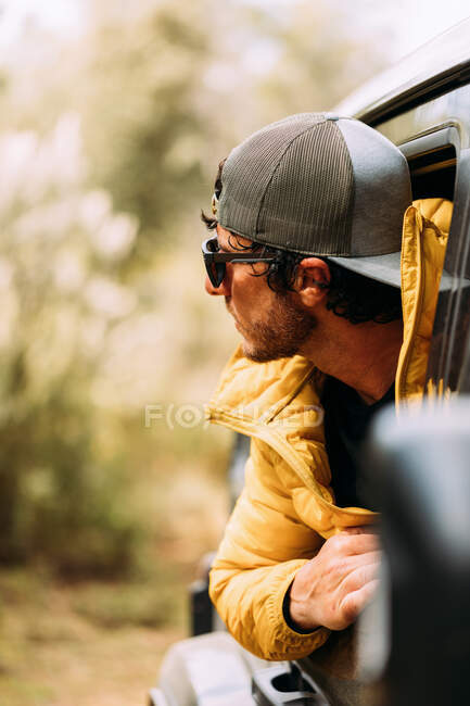 Vista lateral de um aventureiro em um boné e óculos de sol espreitando para fora da venda de carros com fundo borrado — Fotografia de Stock