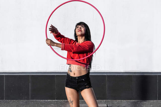 Jeune femme tatouée en vêtements de sport tourbillonnant hula hoop tout en dansant contre des murs de briques avec des ombres et regardant vers l'avant dans la lumière du soleil — Photo de stock