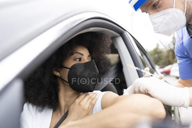 Vista lateral del médico masculino en uniforme protector, guantes de látex y protector facial vacunando a la paciente afroamericana dentro del automóvil en una unidad a través de una clínica móvil durante el brote de coronavirus - foto de stock