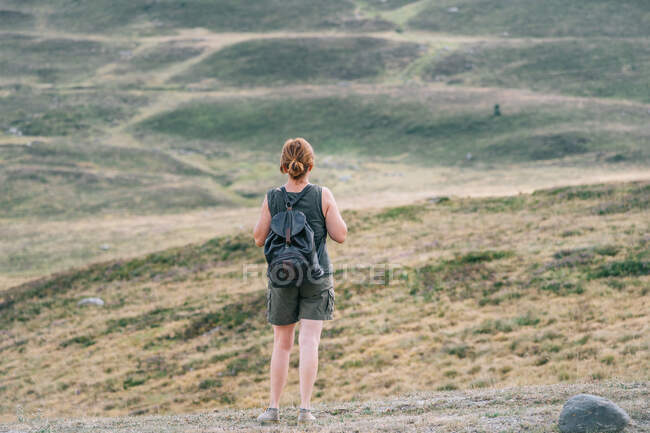 Back view of anonymous female hiker with backpack standing on hill in mountainous terrain and admiring picturesque landscape — Stock Photo