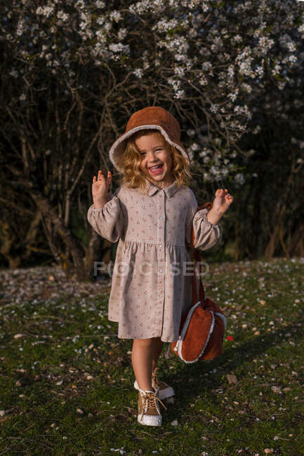 Entzücktes kleines Mädchen in Kleid steht im Frühlingsgarten mit blühenden Blumen auf Bäumen und blickt in die Kamera, während sie den sonnigen Tag genießt — Stockfoto