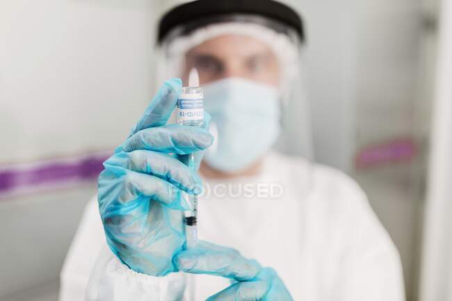 Médico borroso irreconocible con máscara protectora y guantes de látex con vial de vacuna contra el coronavirus y jeringa que se muestra a la cámara mientras está de pie en la habitación del hospital - foto de stock