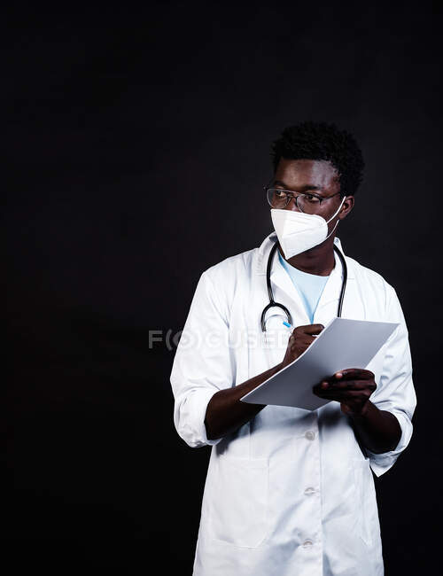 Afro-americano medico maschio in maschera protettiva e uniforme bianca prendere appunti sugli appunti mentre guardando lontano su sfondo nero — Foto stock