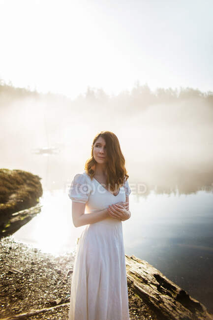 Mujer de pie vestida con un vestido blanco en una roca mirando a un lago en un día de niebla - foto de stock