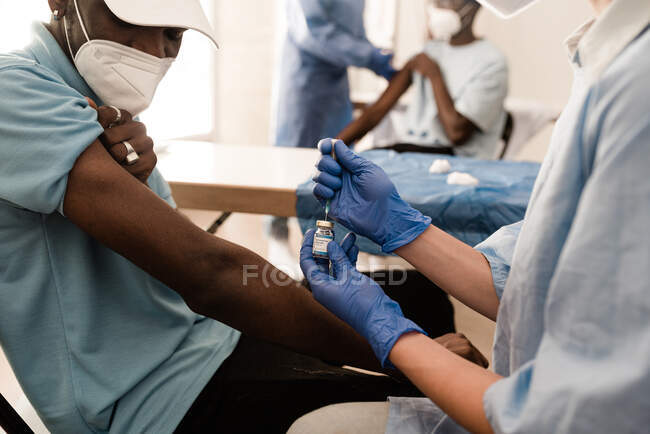 Mãos de corte de médico anônimo em luvas de látex preenchendo seringa de frasco com vacina preparando-se para vacinar paciente afro-americano masculino irreconhecível na clínica durante surto de coronavírus — Fotografia de Stock