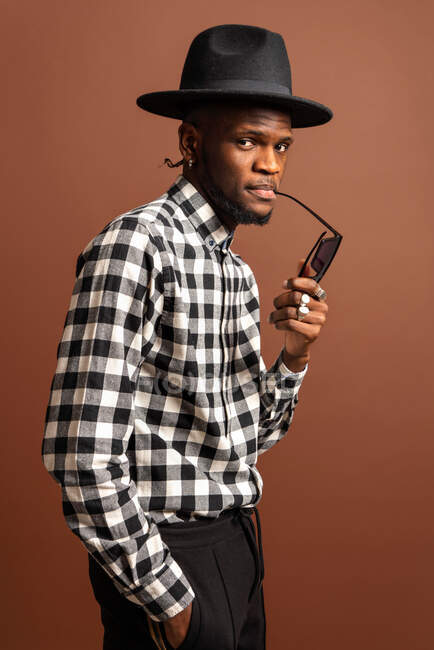 Jovem modelo masculino afro-americano legal em camisa quadriculada e chapéu olhando para a câmera enquanto sentado em fundo marrom — Fotografia de Stock