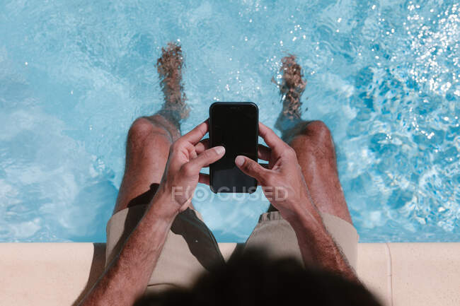 Вид зверху обрізаний невпізнаваний чоловік сидить біля басейну з ногами у воді і переглядає мобільний телефон влітку — стокове фото