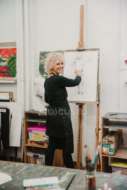 Female artist creating drawing of human with pencil while standing at easel in studio — Stock Photo