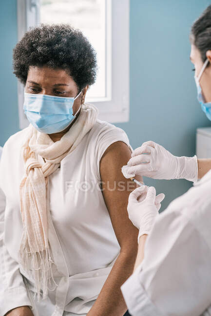 Femme médecin spécialiste en uniforme de protection, gants en latex et masque facial vaccinant femme mûre afro-américaine patiente en clinique pendant l'épidémie de coronavirus — Photo de stock