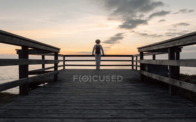 Uomo con zaino e cappello in piedi su una passerella a guardare il mare — Foto stock