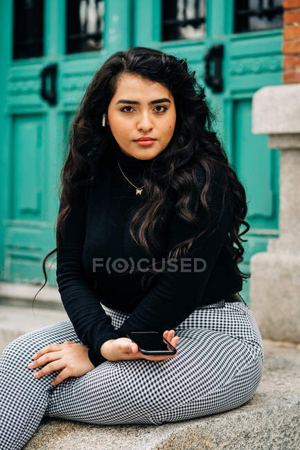 Неэмоциональный плюс размер женщины в модном наряде сидит на каменной границе в городе с мобильным телефоном — стоковое фото