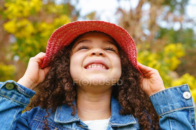 Joyeux enfant en chapeau de paille souriant sur la prairie contre la floraison buisson en plein jour — Photo de stock