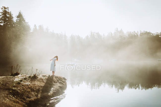 Жінка, одягнена в білу сукню на камені, дивиться на озеро в туманний день. — стокове фото