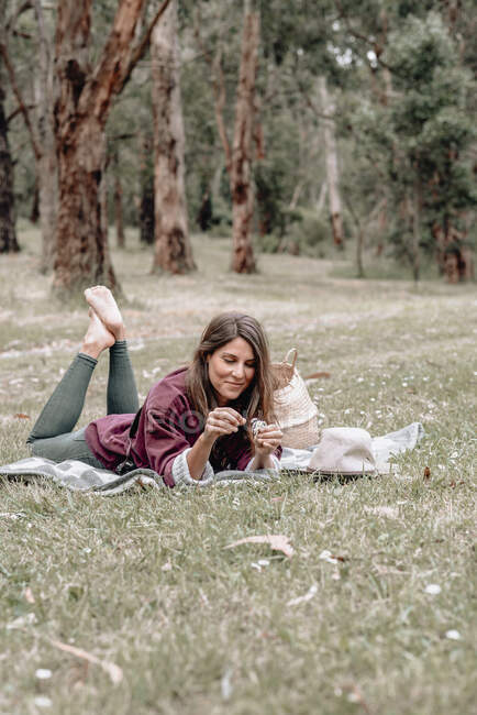 Mulher despreocupada deitada em cobertor na floresta e brincando com a flor margarida enquanto desfruta de piquenique na Austrália — Fotografia de Stock