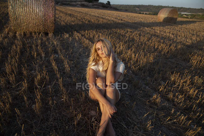 Мирная женщина в элегантном платье сидит на сухом поле в сельской местности и смотрит в камеру — стоковое фото