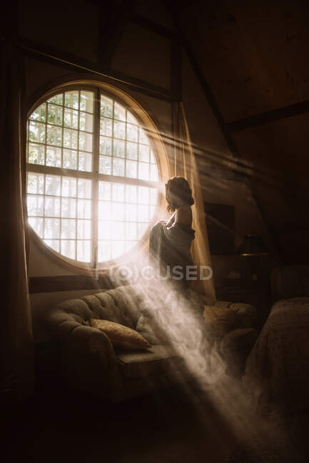 Вид сбоку неузнаваемой женщины в платье, стоящей на диване у круглого окна в доме в солнечный день — стоковое фото