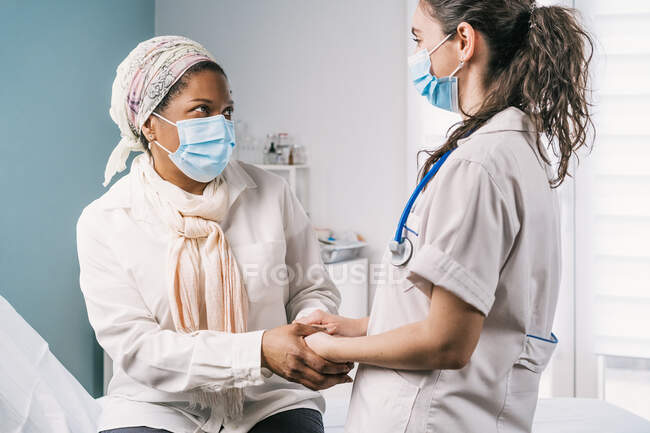 Doctora joven en uniforme médico y estetoscopio que usa mascarilla facial que habla y toma de la mano de una mujer madura afroamericana durante la cita en la clínica - foto de stock
